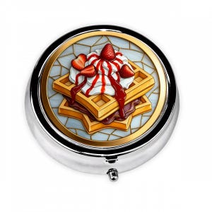 May include: A round, silver pill box with a detailed image of waffles, whipped cream, and strawberries. The waffle design is golden brown, topped with white whipped cream, red strawberry sauce, and sliced strawberries. The image is set against a light blue background.