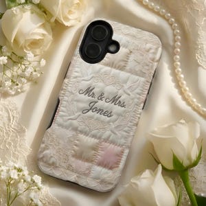 May include: A white and pink quilted phone case with the text "Mr. & Mrs. Jones" embroidered in gray. The case features a lace design and a black camera cutout. The background includes white roses and a pearl necklace.