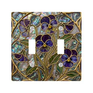 May include: A decorative double light switch cover featuring a floral design with purple pansies and green leaves. The design is set against an iridescent background with gold-colored metal accents. The cover has two switch openings.