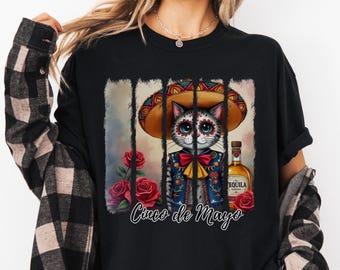 May include: Black t-shirt with a colorful illustration of a cat wearing a sombrero and a traditional Mexican outfit. The cat is holding a bottle of tequila and there are roses and the text "Cinco de Mayo" on the shirt.