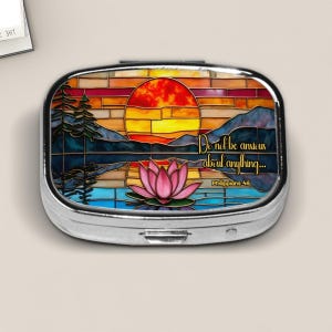 May include: A silver pill box with a stained glass design featuring a sunset over water with a pink lotus flower. The text "Do not be anxious about anything..." is displayed on the box.