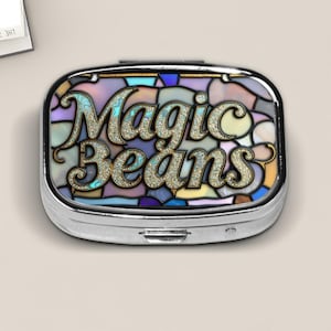 May include: A silver pill box with a colorful stained-glass design. The box features the words "Magic Beans" in a glittery font. The background of the box is a mosaic of pastel colors, including blue, purple, and yellow.