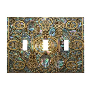 May include: A decorative light switch cover featuring a steampunk design. The cover has a gold-colored frame with gears and spoons. The background has a pearlescent, iridescent finish. The cover has three white switch openings.