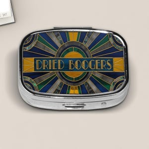 May include: A silver pill box with a colorful Art Deco design. The design features a sunburst pattern in shades of blue, green, and gold, with the words "DRIED BOOGERS" in a blue rectangular banner.