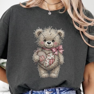 Könnte beinhalten: Dunkelgraues T-Shirt mit einer Teddybär-Illustration. Der Bär hat eine rosa Schleife, einen herzförmigen rosa Patch mit Blumen- und Karomustern und ein rosa Band um den Hals. Das Fell des Bären ist hellbraun.