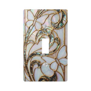 May include: A decorative light switch cover featuring a butterfly and floral design. The cover has a white background with iridescent abalone shell accents and gold-colored metal outlines. The switch opening is white.