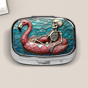 May include: A rectangular silver metal pill case featuring a stained glass style graphic of a white skeleton sitting inside a pink flamingo pool float on blue water. The design has a mosaic appearance with dark outlines and vibrant colors.