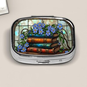 May include: A silver pill box with a stained-glass design featuring a stack of books and blue flowers. The books are in shades of blue, orange, and red. The background has a light blue and white pattern.
