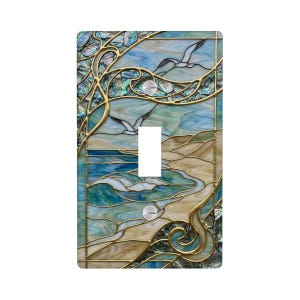 May include: Decorative light switch cover featuring a stained glass design with a coastal theme. The artwork depicts seagulls in flight over a beach and ocean scene, with shades of blue, green, and tan. The design is framed with gold-colored metal accents.