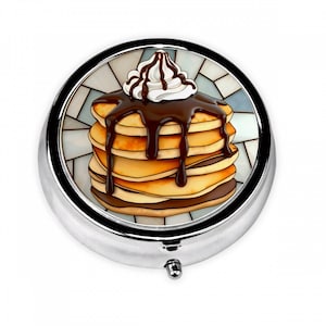 May include: A round, silver pill box with a colorful image of pancakes topped with chocolate syrup and whipped cream. The image is set against a mosaic-style background. The pill box has a small, silver clasp.