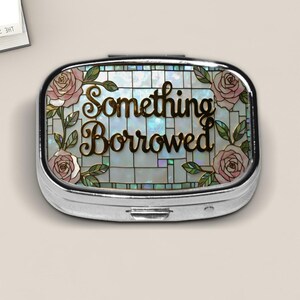 May include: An oval silver-toned metal pill box featuring a decorative top with a stained glass effect. The design includes pink roses with green leaves and the text Something Borrowed in gold script lettering over a pearlescent grid background.