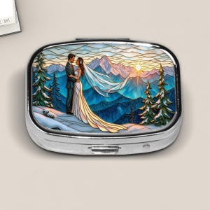 May include: A silver pill box with a mountain scene featuring a couple in wedding attire. The artwork depicts a bride and groom embracing, with a sunset over snow-covered mountains and evergreen trees.