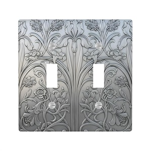 May include: A silver-colored double light switch cover with an Art Nouveau floral design. The cover features raised relief detailing of stylized flowers, leaves, and vines. The two switch openings are rectangular.