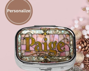 May include: A silver pill box with a stained glass design featuring the name "Paige" in gold lettering. The background of the pill box is a mosaic of pink, white, and gold pieces. The word "Personalize" is in a brown circle.