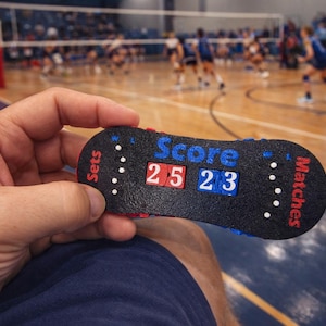 Puede incluir: Un marcador de voleibol negro muestra el marcador 25-23. El dispositivo se sostiene en la mano de una persona, con la cancha de voleibol y los jugadores en el fondo. El marcador tiene detalles en rojo y azul.