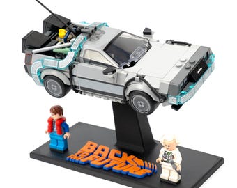 May include: A Lego DeLorean time machine model, gray and white, with light blue accents, is displayed on a black stand. Two Lego figures stand on the base, with the words "BACK TO THE FUTURE" in orange and blue.