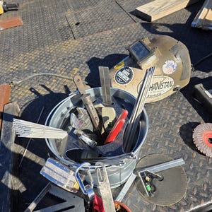 Pu&ograve; includere: Una collezione di strumenti per la saldatura, tra cui un casco per saldatura con adesivi, un secchio pieno di vari strumenti e un disco abrasivo. Gli strumenti sono disposti su una superficie metallica strutturata.