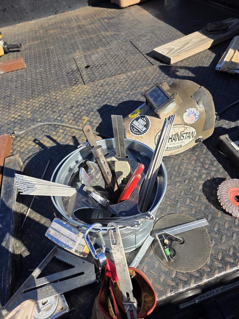 Pu&ograve; includere: Una collezione di strumenti per la saldatura, tra cui un casco per saldatura con adesivi, un secchio pieno di vari strumenti e un disco abrasivo. Gli strumenti sono disposti su una superficie metallica strutturata.