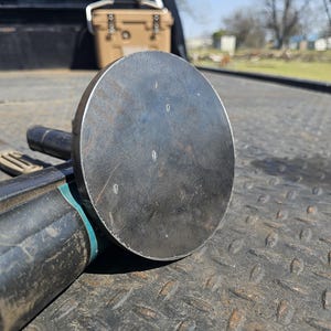 May include: A heavy-duty, circular metal tool with a dark, weathered finish. The tool has a large, flat, disc-shaped head and a cylindrical handle. The head has a few small markings. The tool is likely used for construction or metalworking.