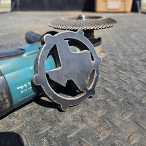 May include: A teal and black angle grinder with a metal Texas-shaped cutting disc attached. The grinder is sitting on a black textured surface.