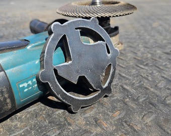 Puede incluir: Una amoladora angular de color azul verdoso y negro con un disco de corte de metal en forma de Texas. La amoladora está sobre una superficie negra texturizada.