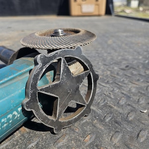 May include: A close-up of a metal star-shaped cutting tool attached to a blue and black angle grinder. The tool is sitting on a textured metal surface.