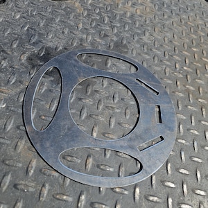 Pu&ograve; includere: Un oggetto metallico circolare con tre grandi fori ovali e diversi fori rettangolari pi&ugrave; piccoli. L'oggetto &egrave; in metallo scuro e poggia su una superficie metallica strutturata. Un altro pezzo metallico circolare &egrave; visibile sullo sfondo.