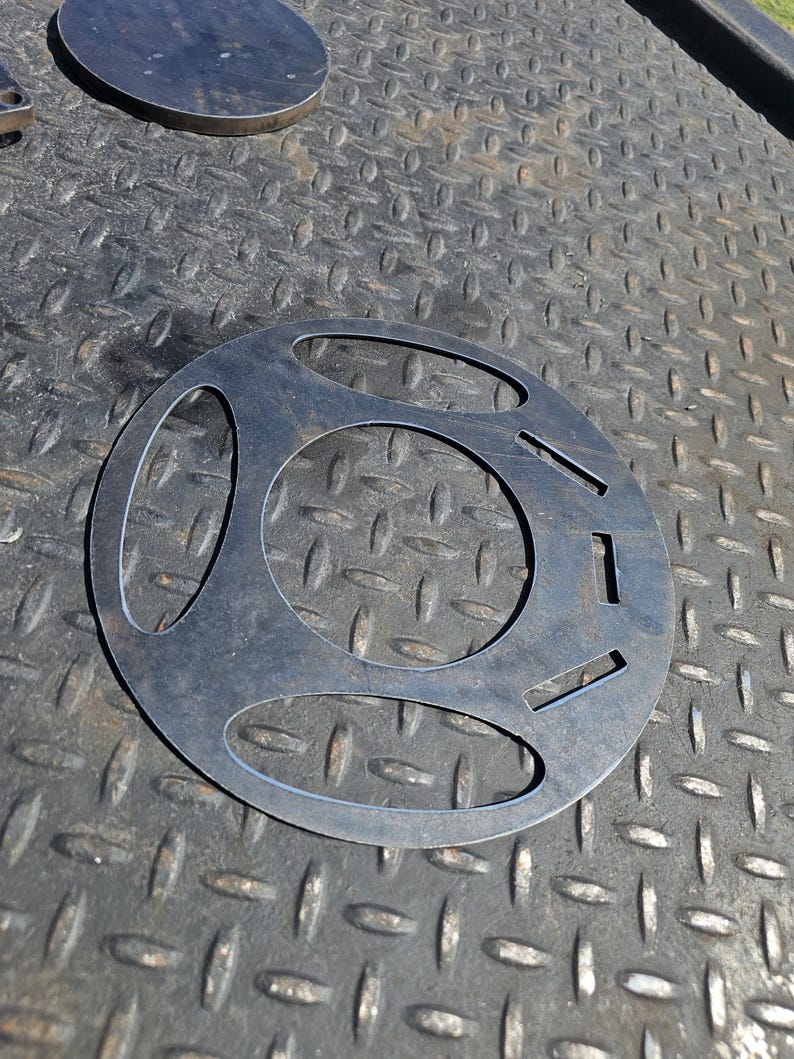 Pu&ograve; includere: Un oggetto metallico circolare con tre grandi fori ovali e diversi fori rettangolari pi&ugrave; piccoli. L'oggetto &egrave; in metallo scuro e poggia su una superficie metallica strutturata. Un altro pezzo metallico circolare &egrave; visibile sullo sfondo.