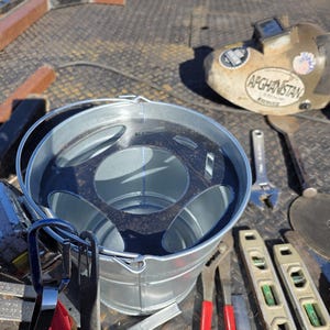 Pu&ograve; includere: Un secchio di metallo zincato con interno scuro e manico, circondato da vari attrezzi. &Egrave; visibile una maschera per saldatura con la scritta "AFGHANISTAN" e adesivi. Altri strumenti includono pinze, una chiave inglese e livelle.