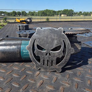 May include: A metal skull-shaped emblem with a circular border, featuring cut-out eyes, nose, and mouth. The emblem is made of weathered metal and rests on a diamond-plate surface. A teal and black power tool is visible.
