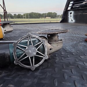May include: A dark teal angle grinder with a circular metal attachment featuring a star-like design with six points and a central hub. The tool is equipped with a circular abrasive flap disc mounted on the top, showing a textured, layered surface.