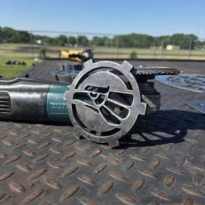 May include: A metal grinder with a circular, gear-shaped attachment featuring a stylized eagle design. The tool is teal and gray, resting on a textured metal surface. Additional grinding discs are visible in the background.