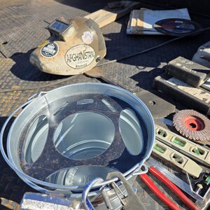 Pu&ograve; includere: Una collezione di strumenti per la lavorazione dei metalli, tra cui un casco per saldatura con un adesivo "Afghanistan Served", un secchio di metallo, livelle e una sega. Gli strumenti sono disposti su una superficie metallica strutturata.