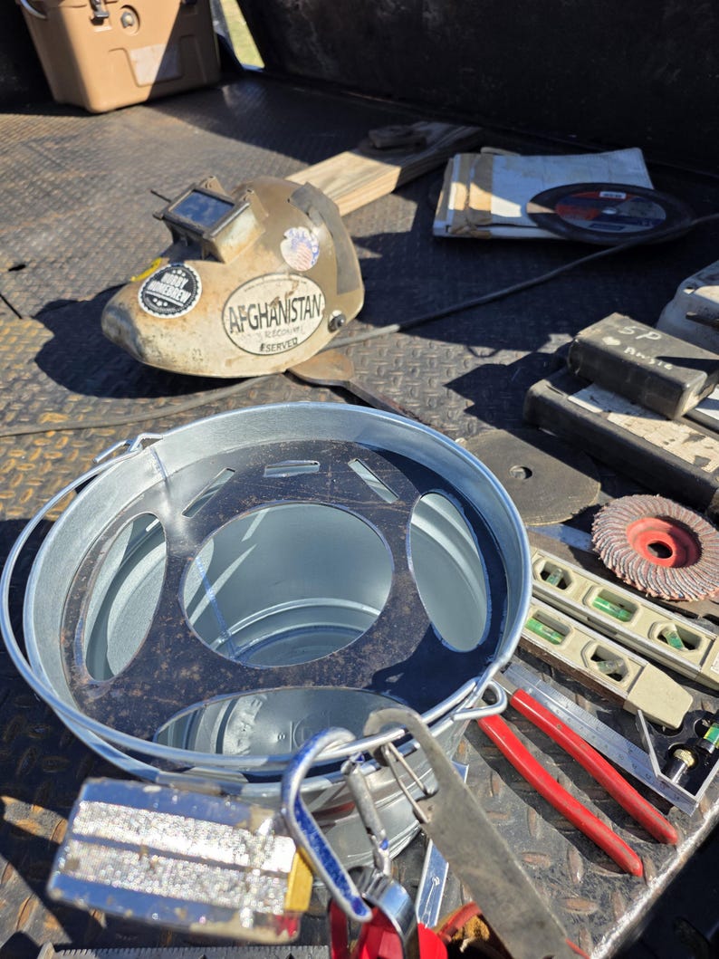Pu&ograve; includere: Una collezione di strumenti per la lavorazione dei metalli, tra cui un casco per saldatura con un adesivo "Afghanistan Served", un secchio di metallo, livelle e una sega. Gli strumenti sono disposti su una superficie metallica strutturata.