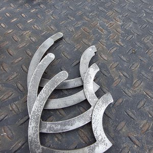 May include: Several silver-colored, curved metal pieces with a distressed finish are arranged on a textured, dark gray surface. The pieces have a unique, open-ended shape, possibly for industrial or artistic use.