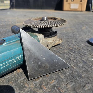 May include: A close-up of a metal angle grinder with a disc and a triangular metal piece. The grinder is teal and silver, and the disc is a dark, circular shape. The metal piece is a dark gray color.