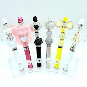 May include: A collection of six decorative pens with various embellishments. The pens have white, pink, black, silver, and yellow barrels. Each pen features a unique topper, including hearts, bows, and floral designs, all adorned with rhinestones and beads. They are displayed on a clear acrylic stand.