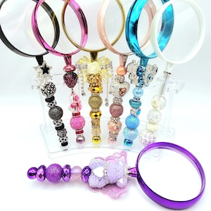 May include: A collection of decorative magnifying glasses with various colored frames and embellished handles. The handles feature beads, stars, and other decorative elements. The frames are black, pink, gold, rose gold, teal, white, and purple.