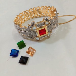 Peut inclure: Un bracelet doré et argenté avec une pierre rouge carrée au centre. Le bracelet présente un motif de filigrane décoratif et est accompagné de quatre pierres carrées lâches bleues, vertes, orange et noires.