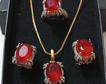 May include: A jewelry set featuring a necklace, ring, and earrings, all with large, faceted red gemstones. The stones are set in ornate, silver-toned metal with gold accents, presented in a black box.