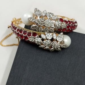 Könnte beinhalten: Zwei goldfarbene Armreifen mit roten und klaren Edelsteinakzenten. Jeder Armreif hat eine große weiße Perle und ein Blumendesign mit klaren Edelsteinen. Die Armreifen liegen auf einer schwarzen Oberfläche.