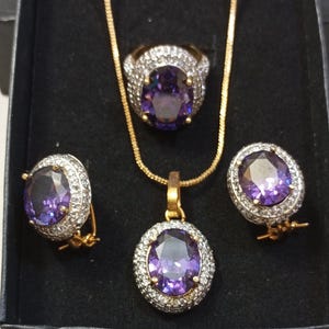以下が含まれることがあります： 金色のネックレス、指輪、イヤリングがセットになったジュエリーセット。セットには、小さな透明な石に囲まれた大きな楕円形の紫色の宝石が付いた指輪とイヤリングが含まれています。ネックレスにはお揃いのペンダントが付いています。