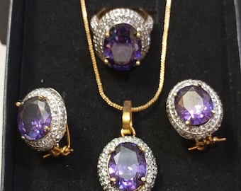 May include: A jewelry set featuring a gold necklace, ring, pendant, and earrings. Each piece has a large, oval, purple gemstone surrounded by small, clear stones. The set is displayed in a black box.