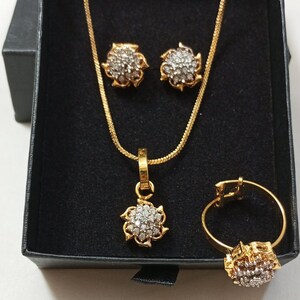 May include: A gold-toned jewelry set consisting of a necklace with a floral-shaped pendant, a pair of matching stud earrings, and a ring. Each piece features a cluster of small, clear, sparkling stones set within a gold-colored metal frame.