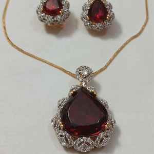Puede incluir: Un collar de oro con un colgante que presenta una gran piedra preciosa roja facetada rodeada de piedras blancas más pequeñas. El collar se combina con pendientes a juego, cada uno con una piedra roja más pequeña rodeada de piedras blancas. El conjunto es un diseño clásico y elegante.