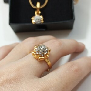 May include: A gold-toned ring featuring a cluster of small clear stones arranged in a floral pattern. In the background, a matching gold-toned necklace with a floral pendant and a pair of matching stud earrings are displayed in a black box.