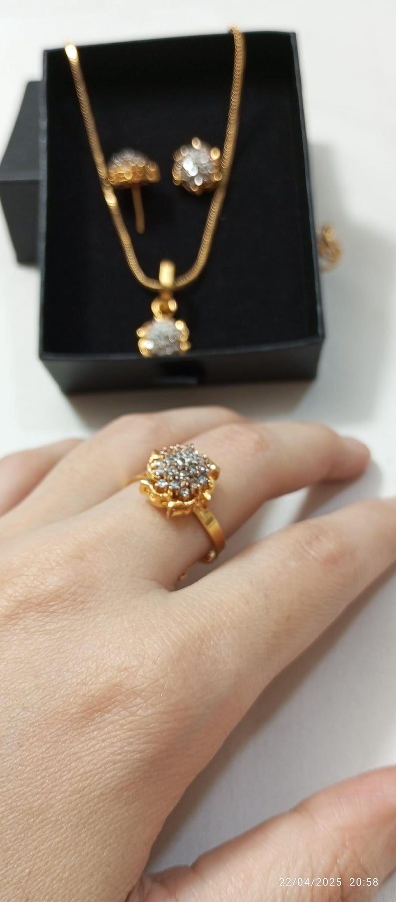 May include: A gold-toned ring featuring a cluster of small clear stones arranged in a floral pattern. In the background, a matching gold-toned necklace with a floral pendant and a pair of matching stud earrings are displayed in a black box.