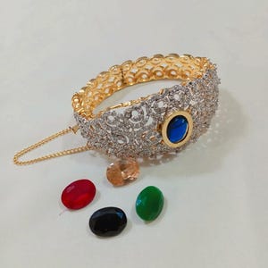 May include: A gold and silver-toned bracelet with intricate detailing and a blue gemstone. The bracelet is accompanied by several colorful gemstones in red, orange, black, and green. A delicate chain is attached to the bracelet.