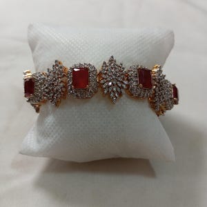 Könnte beinhalten: Ein goldfarbenes Armband mit rechteckigen roten Edelsteinen und funkelnden, klaren Kristallen. Das Armband liegt auf einem weißen Kissen, das sein Design hervorhebt. Die Edelsteine sind von kleineren Kristallen umgeben, was einen funkelnden Effekt erzeugt.