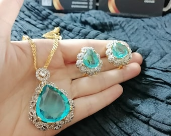 May include: A gold-toned necklace and earring set featuring large, teardrop-shaped teal gemstones. The gemstones are surrounded by a halo of smaller, clear stones. The set includes a pendant necklace and matching stud earrings.